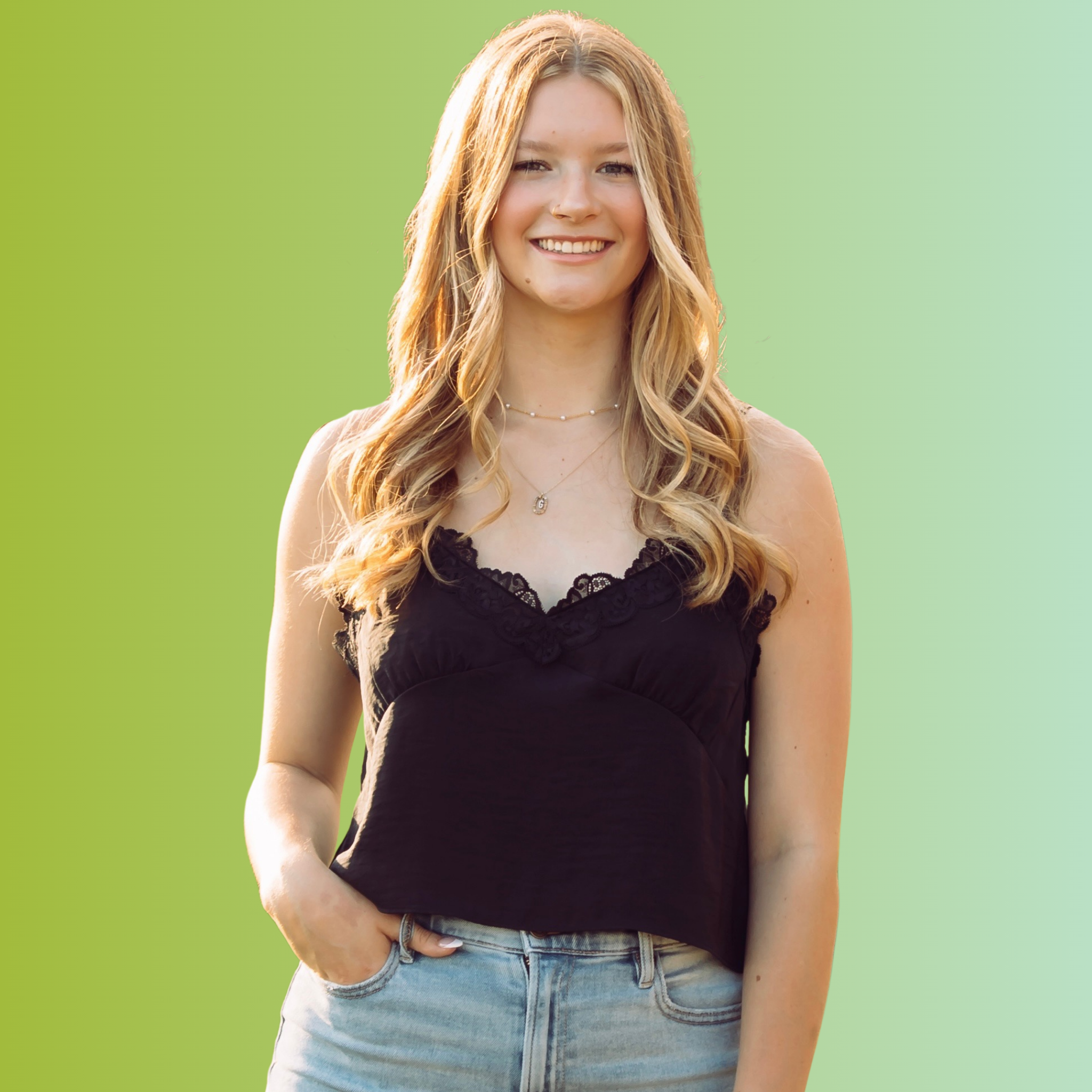 Young woman with long, wavy blonde hair, wearing a black lace-trimmed sleeveless top and light blue jeans, smiling outdoors against a green background.