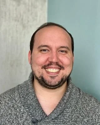 Blake Bettis, a white man in a grey sweater, is standing in front of a blue and grey background. Shown from shoulders up, smiling at camera.