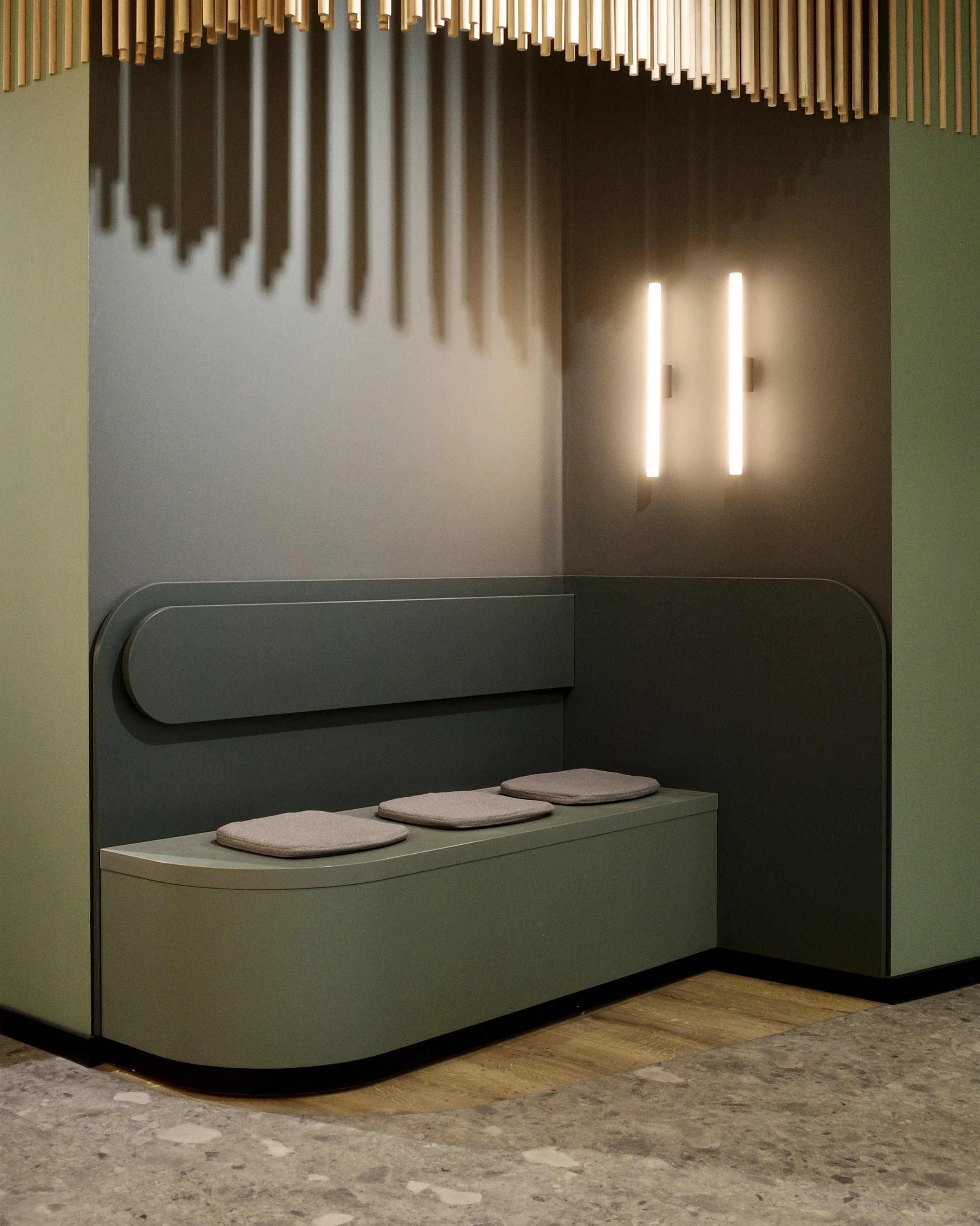 Foto unserer Wartezone mit modernem Sitzbereich mit grüner, gebogener Sitzbank, drei Polsterkissen, dunklen Wänden, vertikalen Lichtleisten, Holzdekor an der Decke.