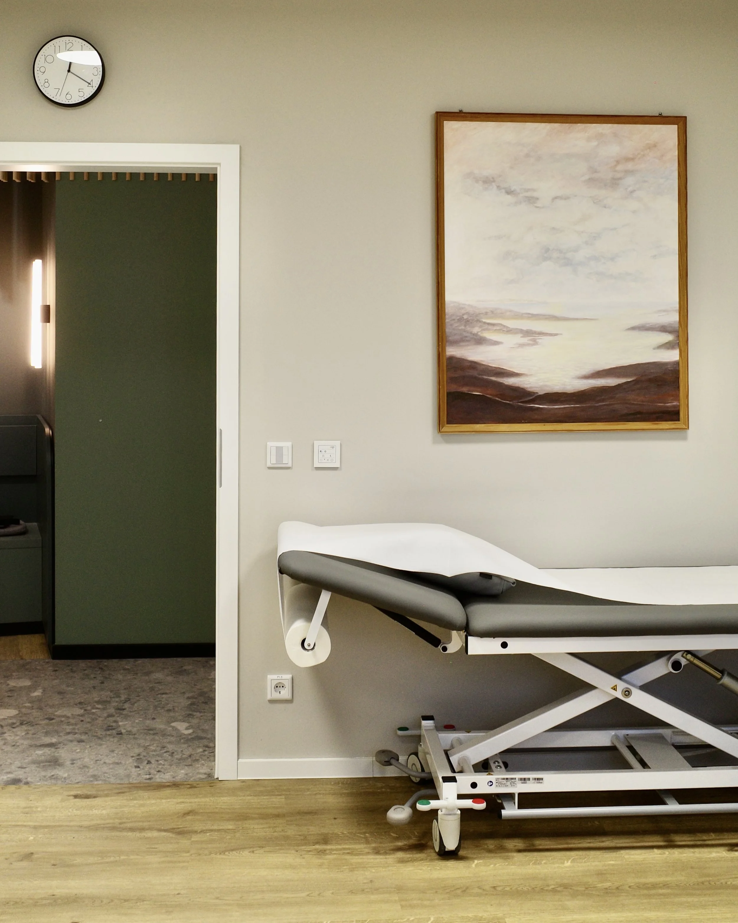 Foto eines Behandlungszimmers mit Untersuchungsliege, einem Panorama-Gemälde an der Wand, einer Uhr, Lichtschaltern und einer Tür zum Flur.