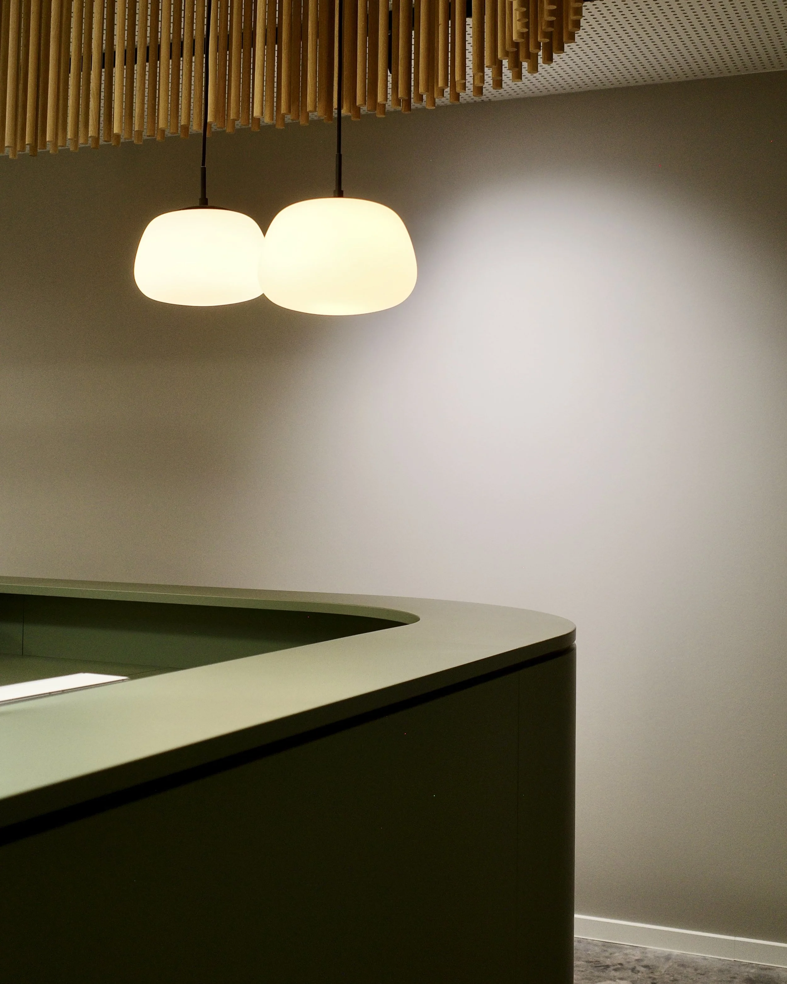 Foto unseres Eingangsbereiches mit einer modernen Lichtinstallation mit zwei weißen Lampenschirmen, hängend über einer grünen Empfangstheke in einem Raum mit weißer Wand und einer Decke mit Holzelementen.