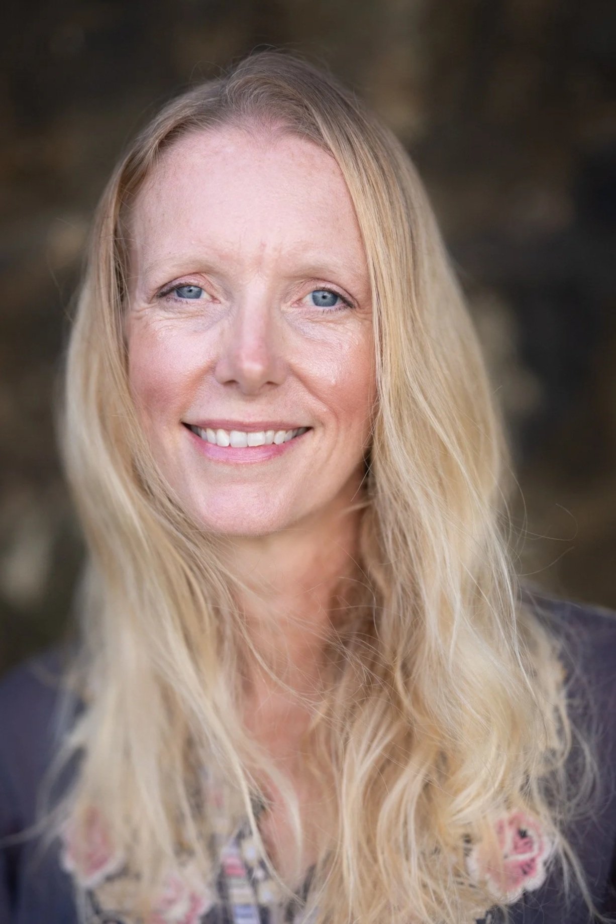Portrait of a woman with long blonde hair, blue eyes, and a friendly smile, outdoors with a blurred natural background.
