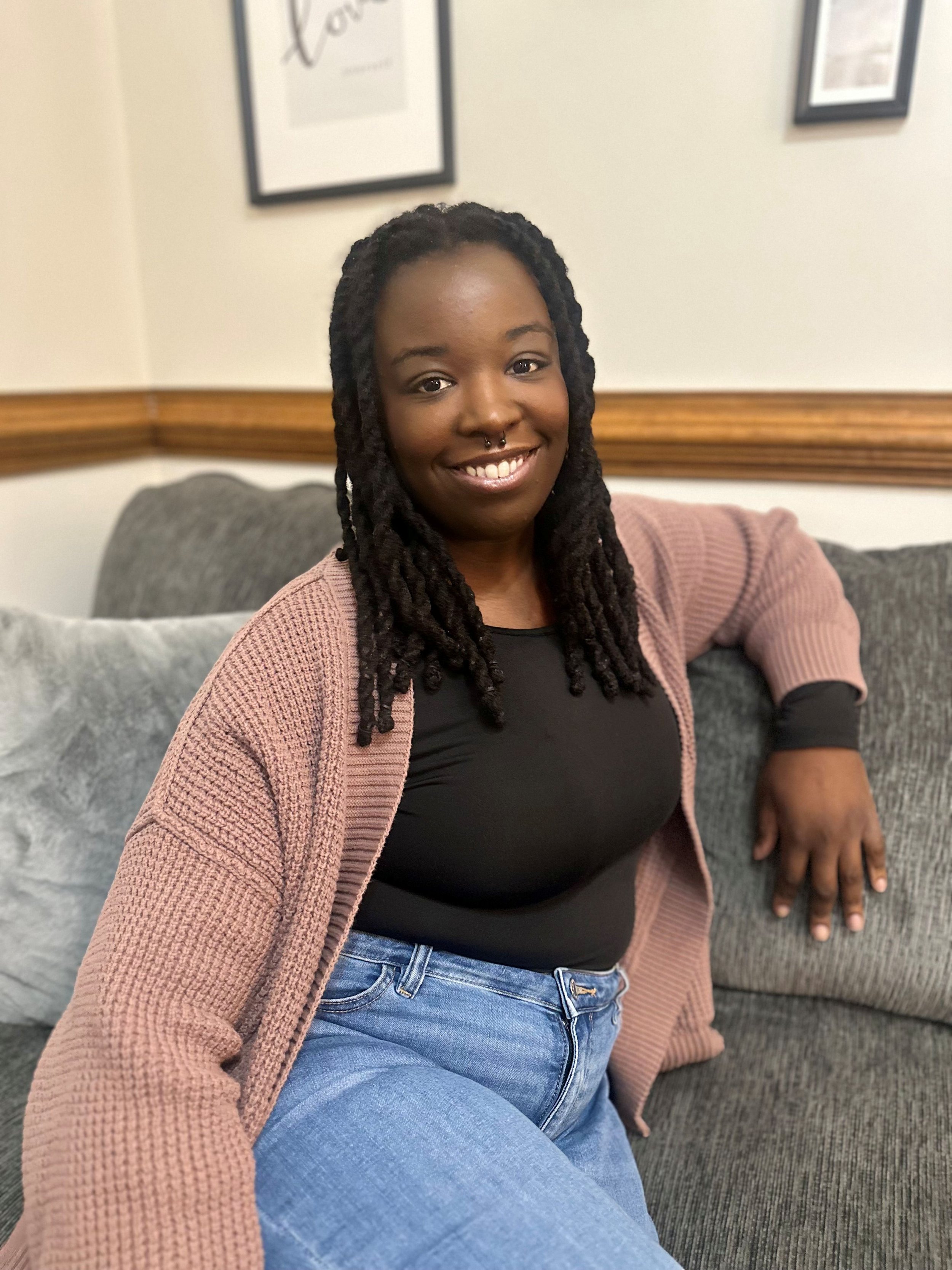 A smiling woman with long dreadlocks sitting on a gray couch in a room with framed pictures on the wall.