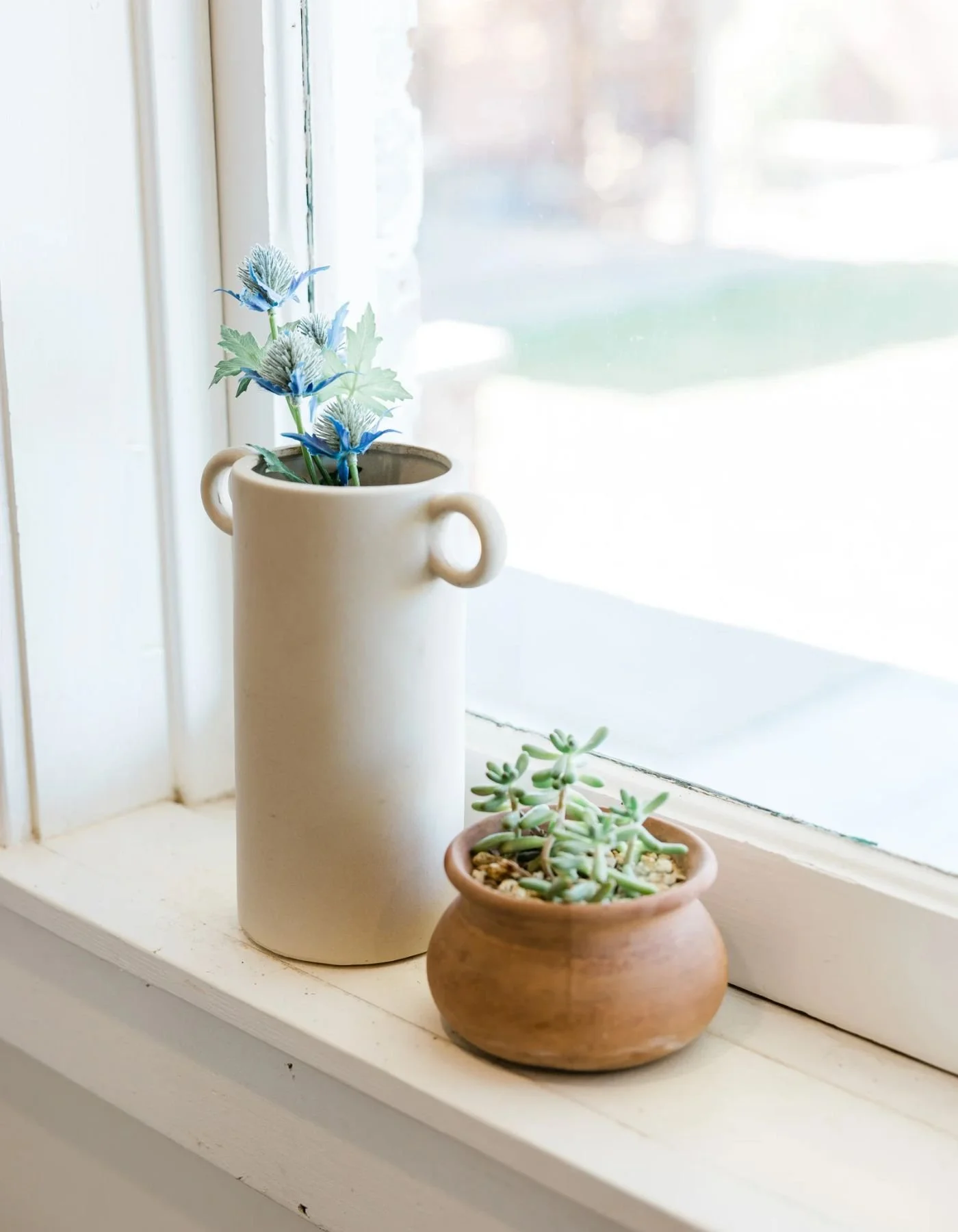 A windowsill with a tall white vase holding blue artificial flowers and a small terracotta pot with green succulent plants.