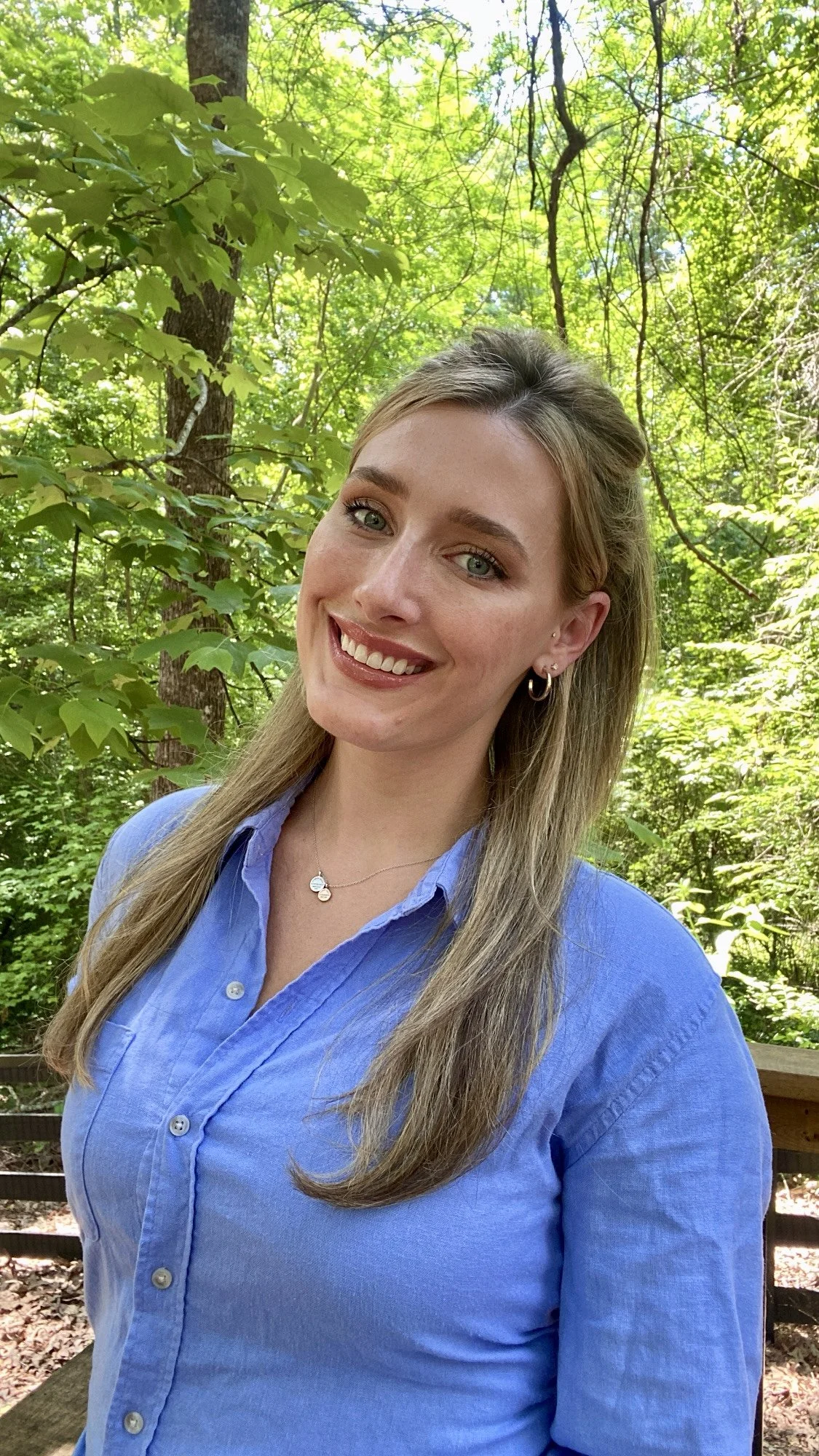 A young woman with light brown hair and blue eyes, wearing a blue button-up shirt, standing outdoors in a lush green forest, smiling at the camera.
