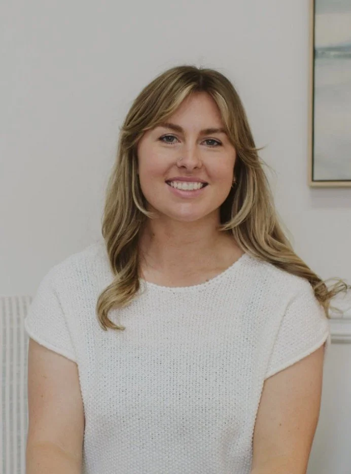 A woman with shoulder-length blonde hair smiling, wearing a white short-sleeve top, standing in front of a plain wall with a framed artwork visible on the side.
