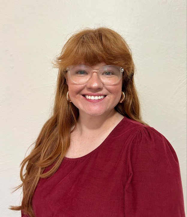 A woman with long red hair, wearing glasses and gold hoop earrings, smiling, dressed in a burgundy top, standing against a plain wall.