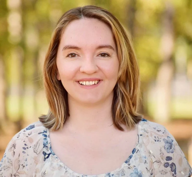 A smiling young woman with shoulder-length light brown hair, wearing a floral top, standing outdoors with trees in the background.