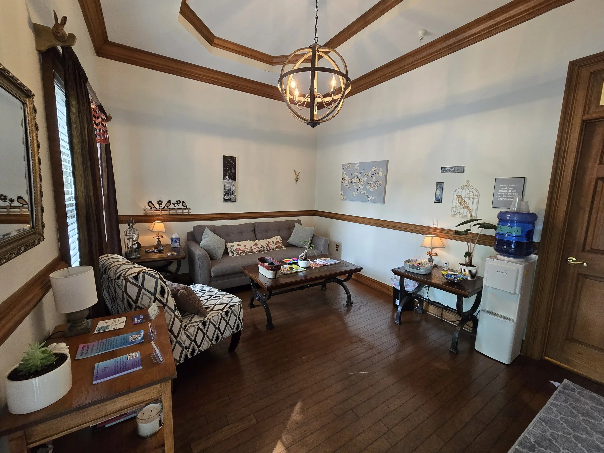 A room with wooden floors, cream walls with wooden trim, a large window with brown curtains, a gray couch with pillows, a patterned armchair, a wooden coffee table, and a side table with a lamp. There are wall decorations including paintings, a birdcage, a small clock, and a water dispenser on a small table.