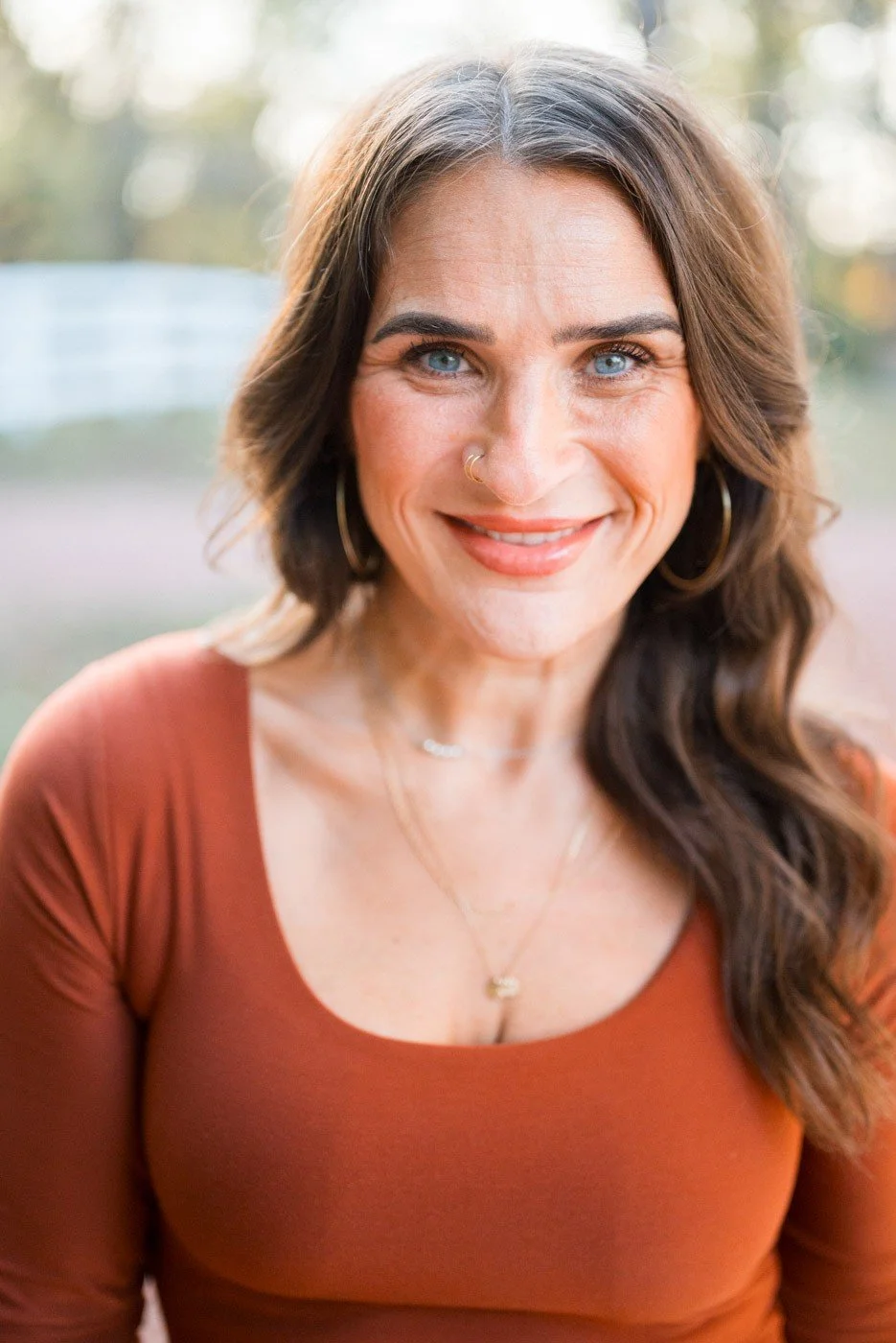 Close-up of a woman with blue eyes, brown wavy hair, wearing a rust-colored top, gold hoop earrings, and layered necklaces, smiling outdoors.