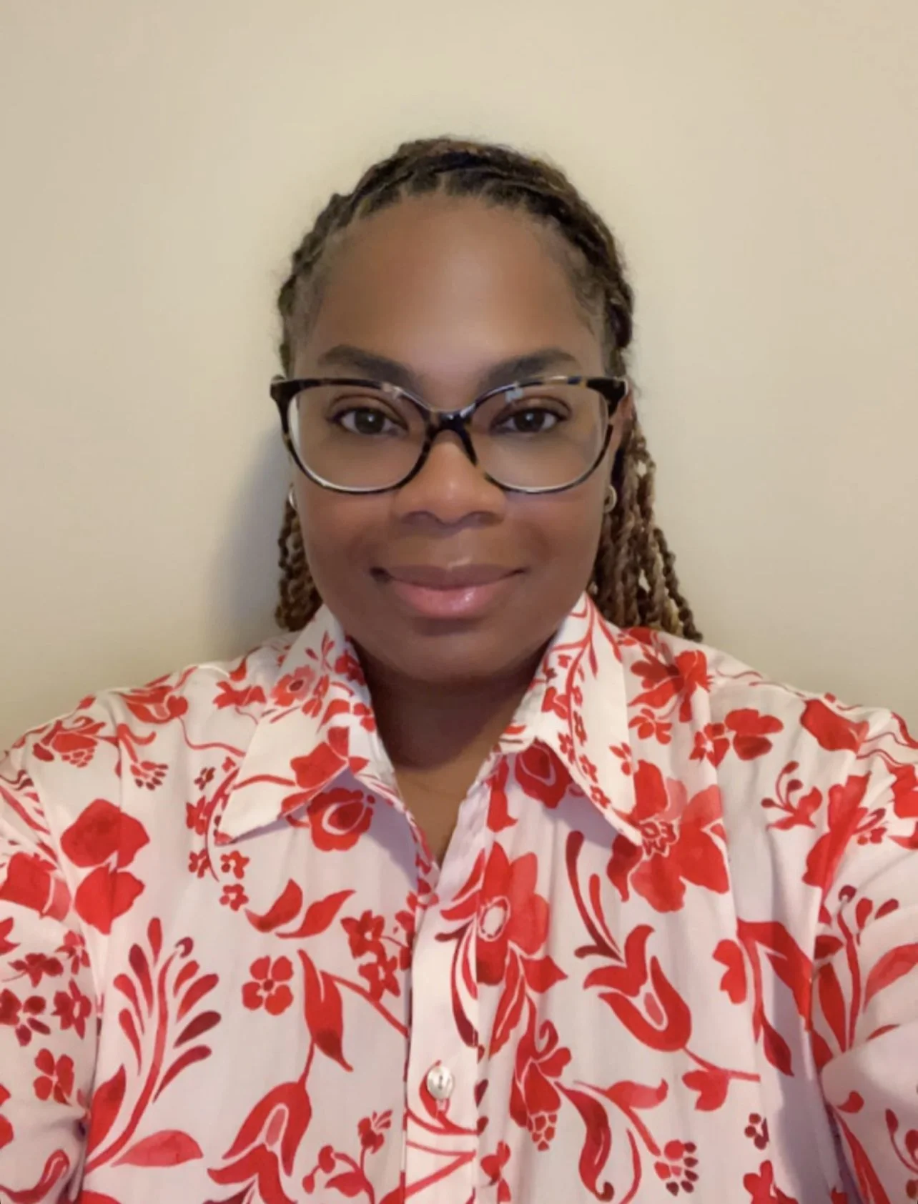 A woman with glasses and braided hair taking a selfie against a plain beige wall, wearing a white shirt with red floral patterns.