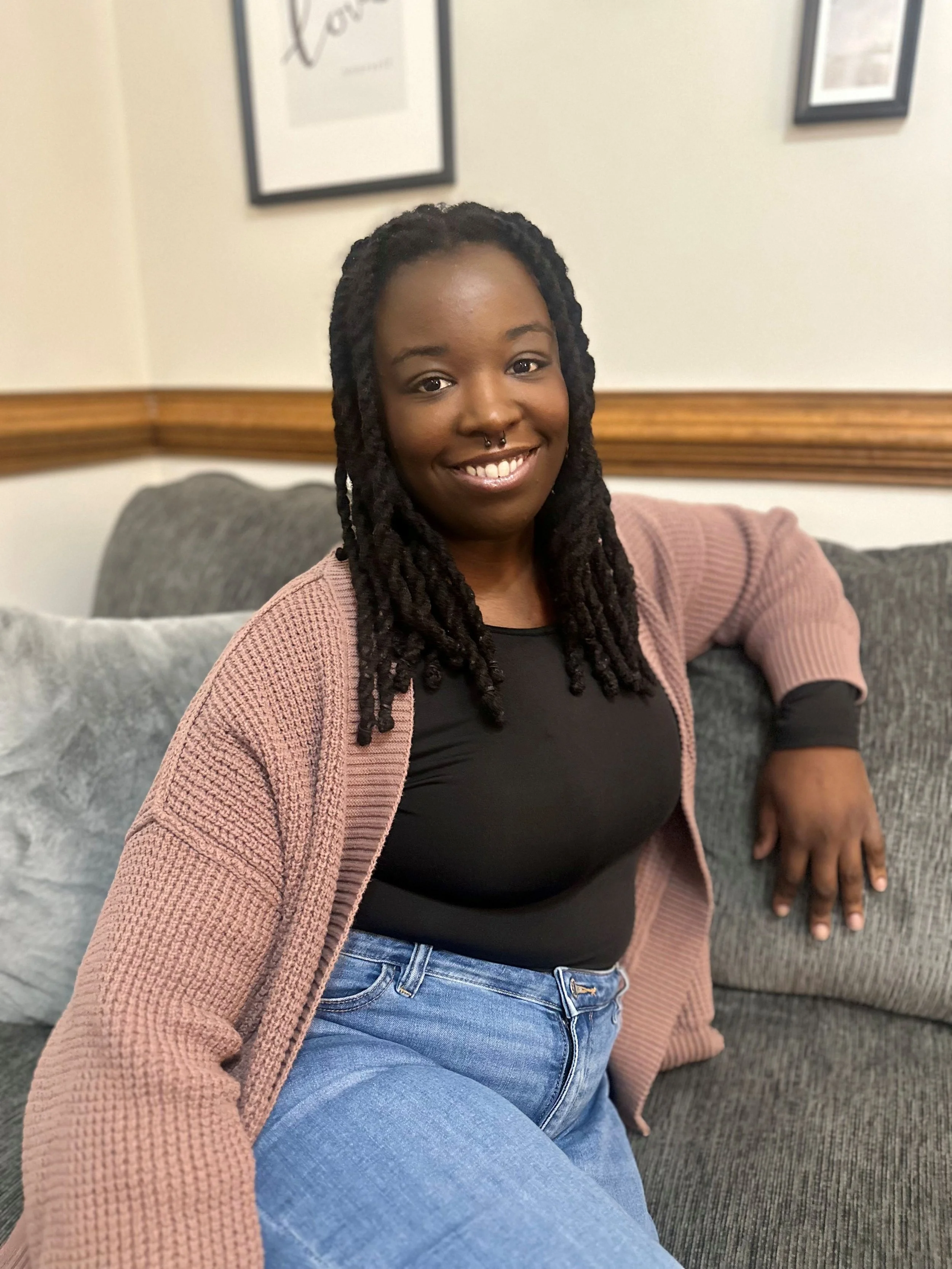 A young woman with dreadlocks, smiling, sitting on a gray couch in a room with framed art on the walls.