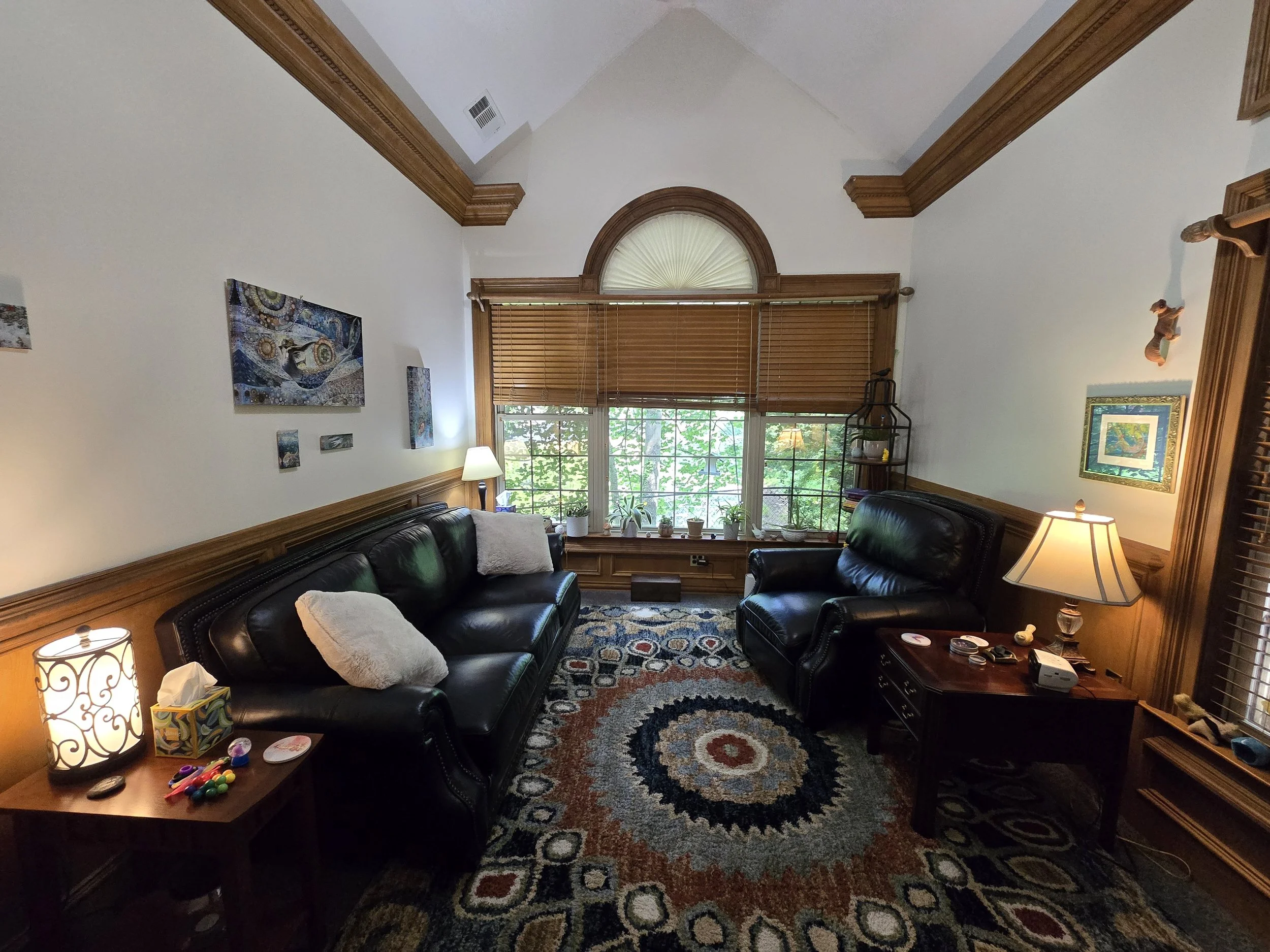 Living room with two black leather sofas, a wooden side table with colorful toys and tissues, a floor lamp, an area rug with circular patterns, large window with blinds, and artwork on the walls.