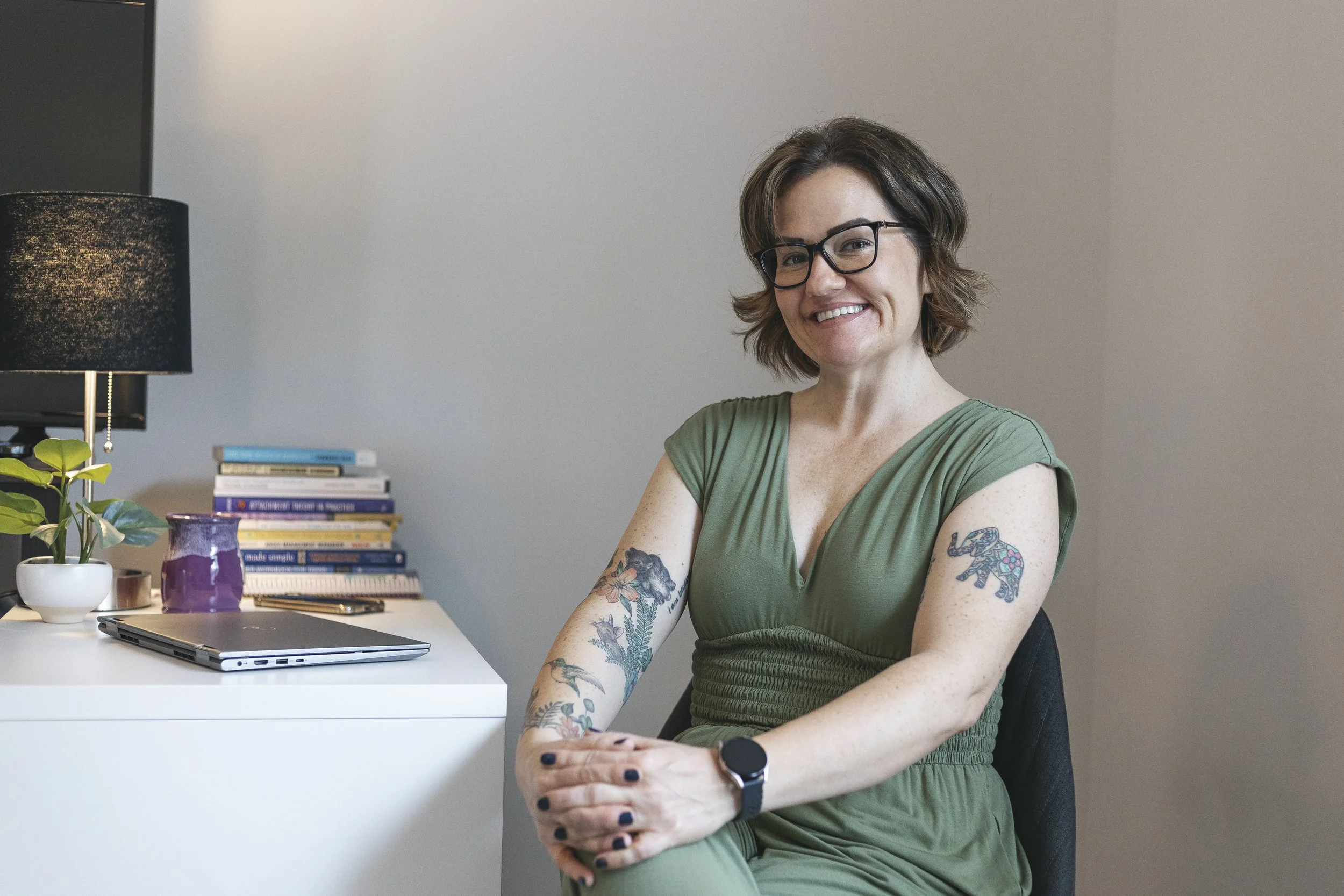 A woman in a green dress sitting in a chair, smiling, with tattoos on her arms, glasses, a smartwatch, and a short haircut, in a room with a desk, books, a plant, and a lamp.