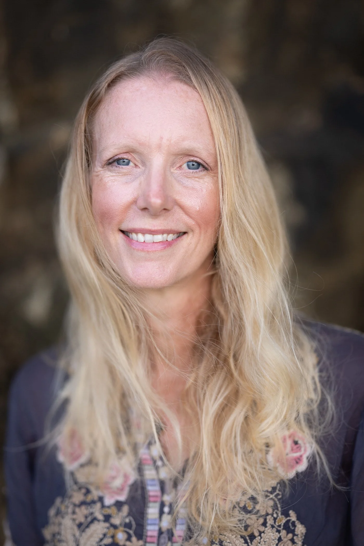 Close-up portrait of a smiling woman with long blonde hair, blue eyes, and light skin, outdoors with blurred natural background.