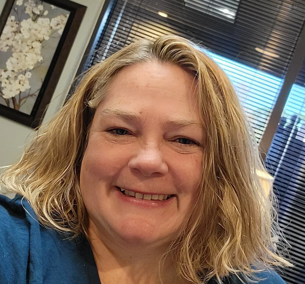 A woman with curly blonde hair, smiling, indoors with window blinds in the background.