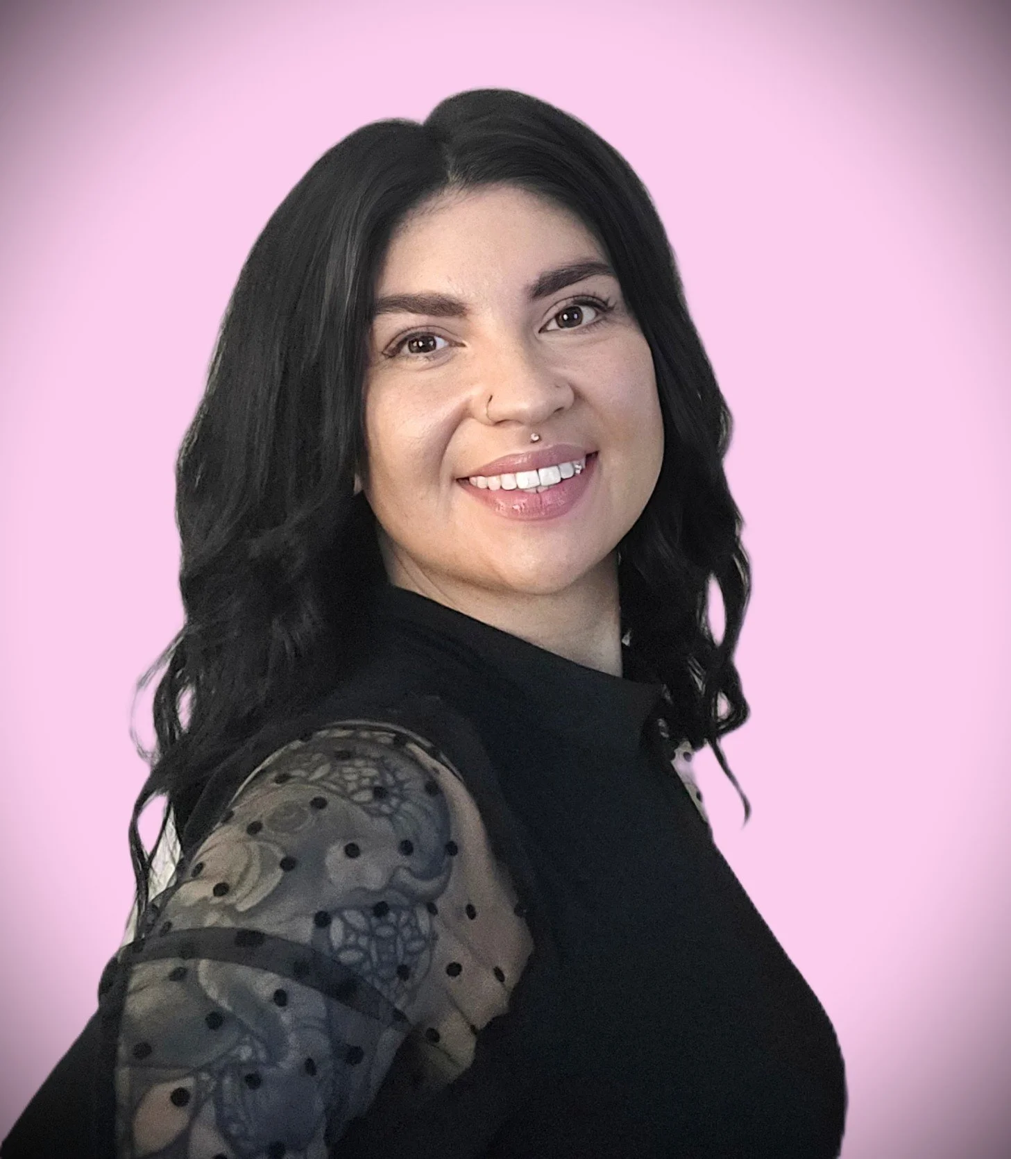 A woman with black hair, a septum piercing, and a smile, wearing a black top with sheer sleeves, standing against a pink background.