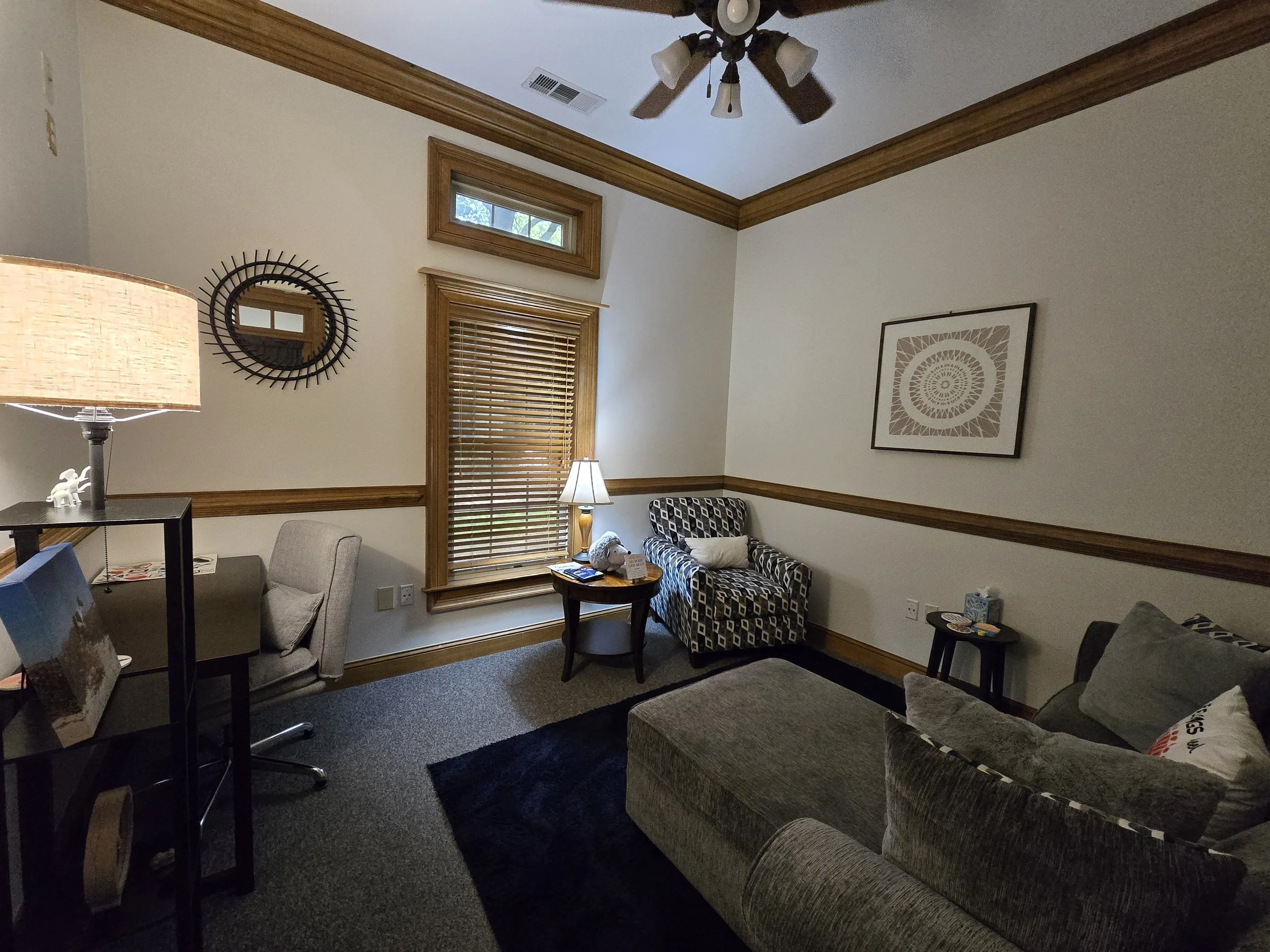 Living room with beige walls, wooden trim, a ceiling fan, a window with blinds, a gray sofa with pillows, a patterned cushioned chair, a wooden side table with a lamp, and framed artwork on the wall.
