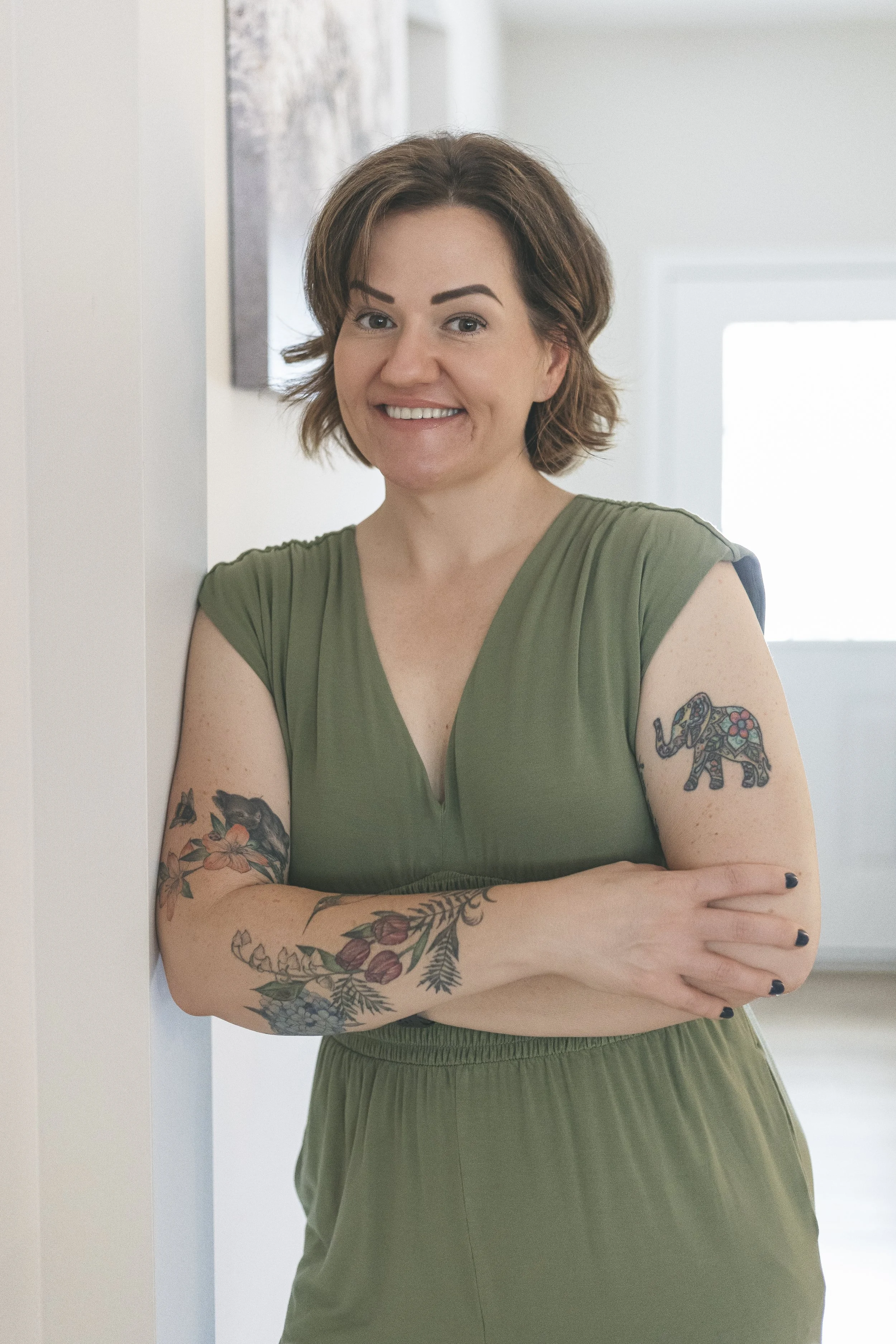 A woman with short brown hair and tattoos on her arms, wearing a green dress, smiling and standing with her arms crossed inside a room.