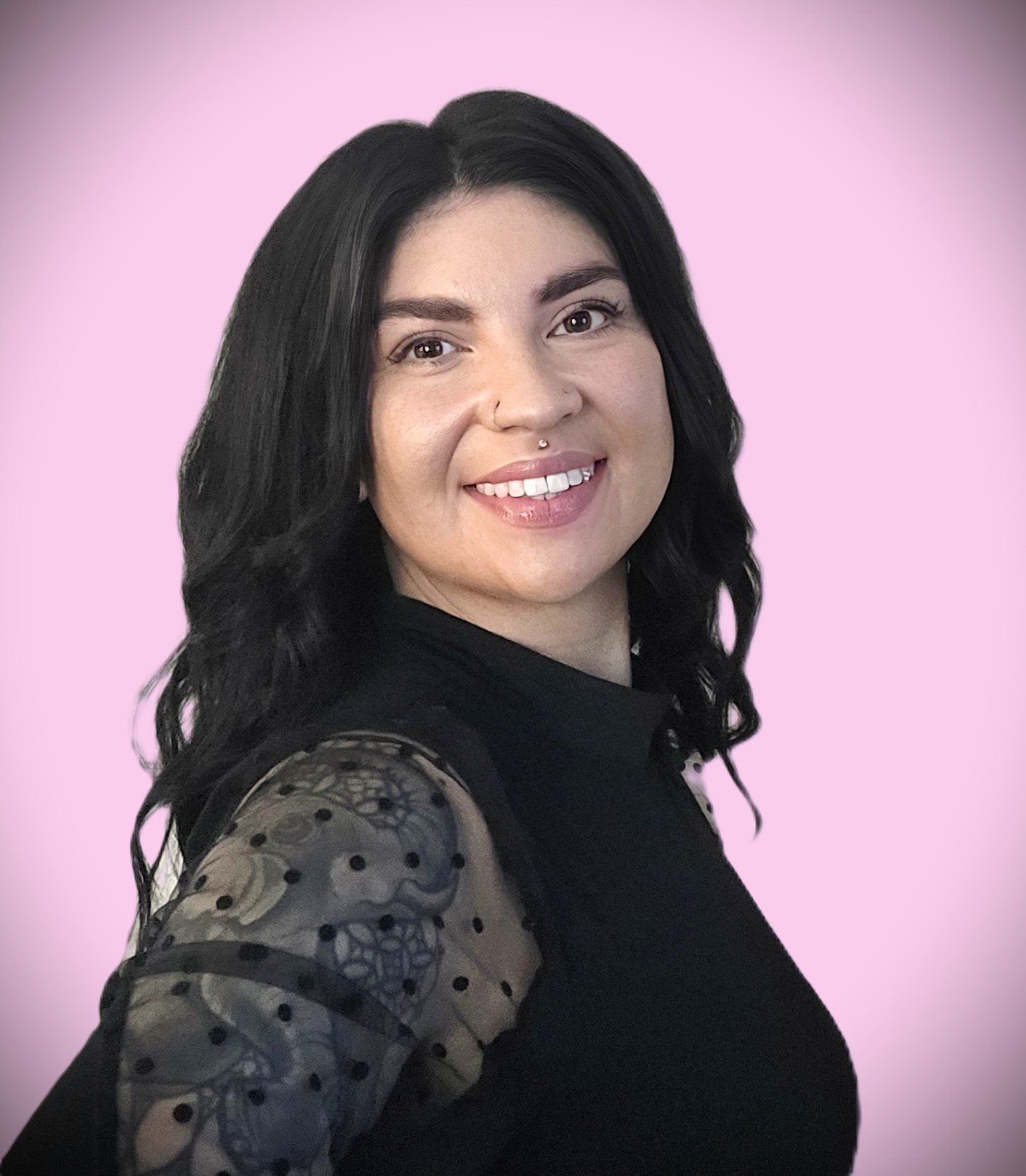 Portrait of a woman with black wavy hair, smiling, wearing a black top with sheer polka dot sleeves, against a pink background.