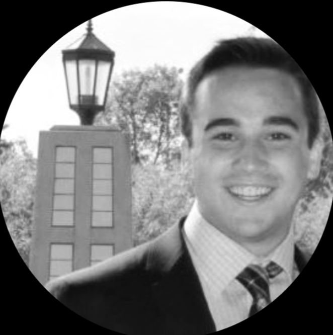 Young man in business suit smiling outdoors with a lamp post and trees in the background.