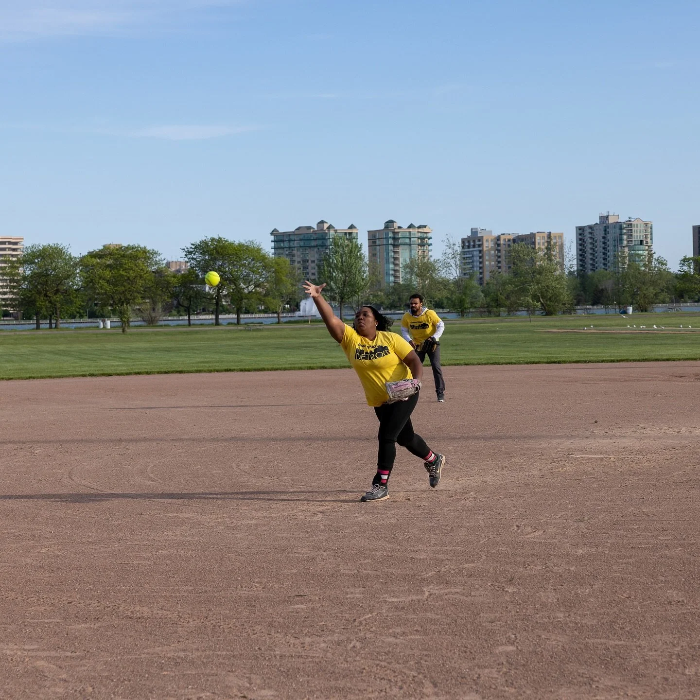 Two people playing softball on a dirt field with green grass and trees, city buildings in the background, one woman in yellow throwing a yellow ball, a man in yellow behind her.