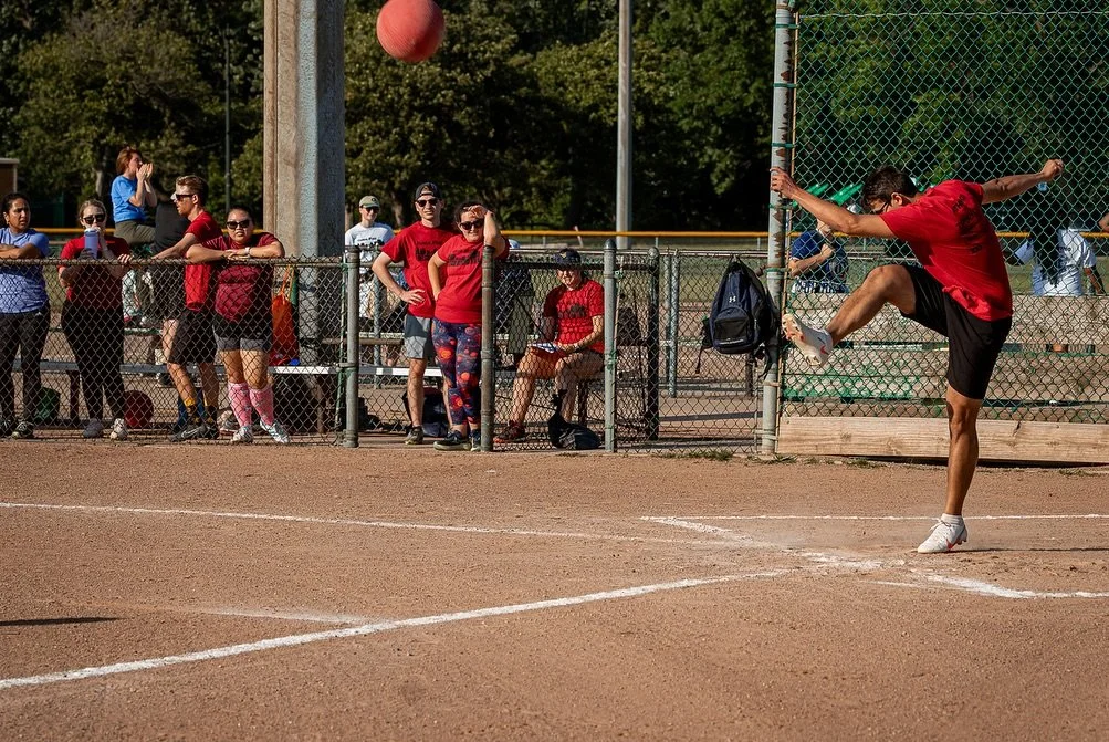 A person in a red shirt and black shorts is kicking a ball on a dirt sports field, with spectators watching from behind a chain-link fence.