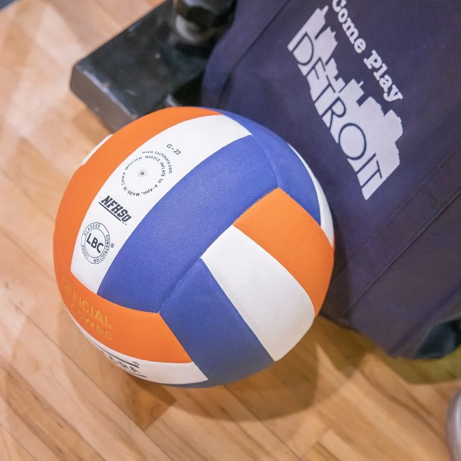 A volleyball with orange, blue, and white panels next to a navy bag with the text 'Come play Detroit' and a city skyline.