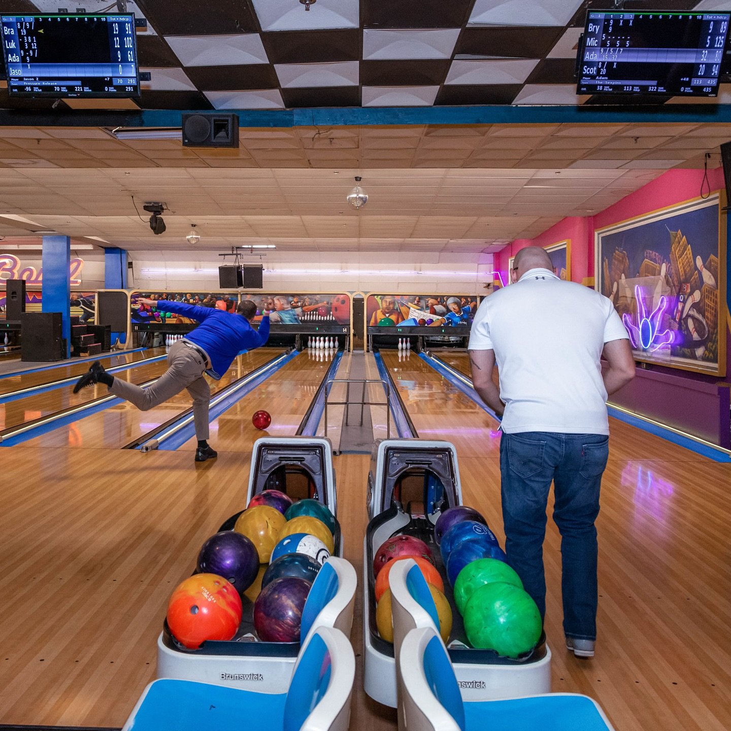 People bowling in a bowling alley with colorful bowling balls in the foreground and two electronic scoreboards hanging from the ceiling.
