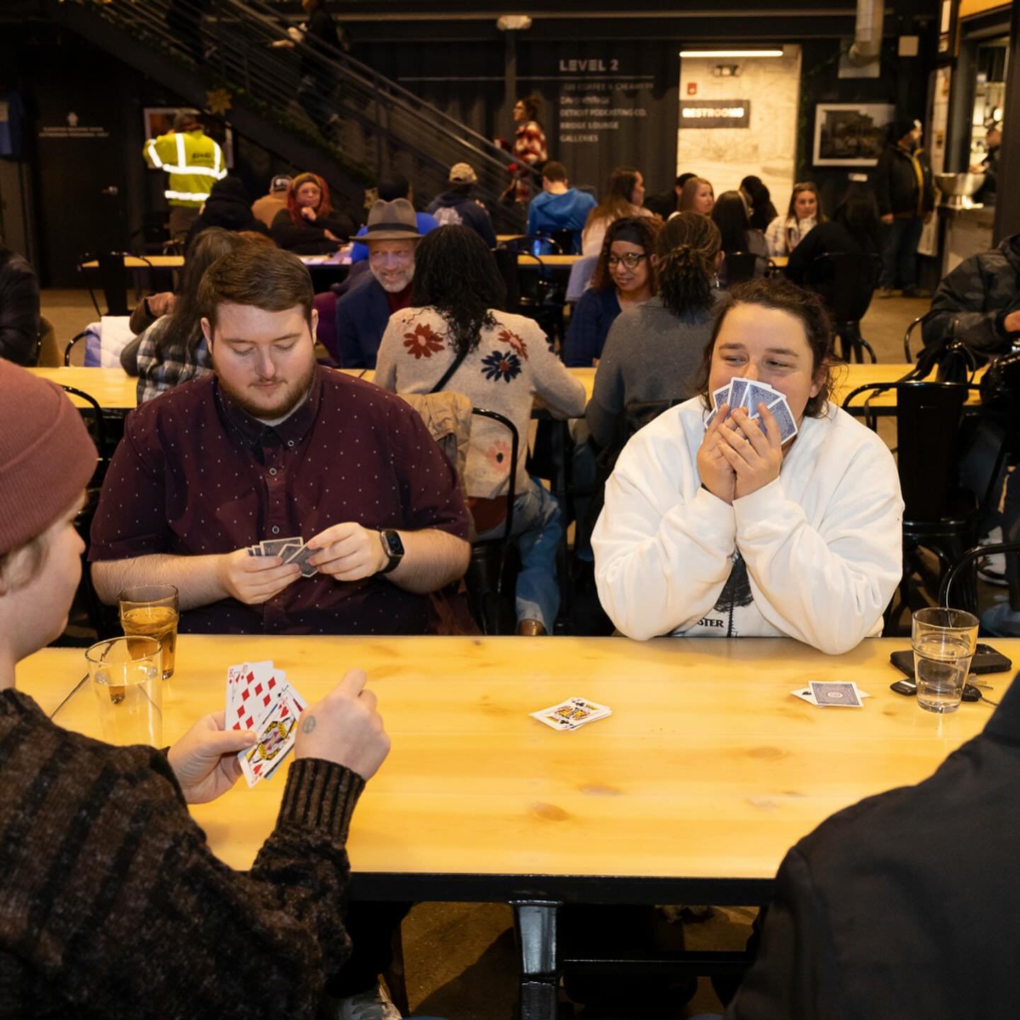 People playing cards at a table in a crowded indoor space, with glasses of drinks on the table and a sign indicating 'Level 2' in the background.