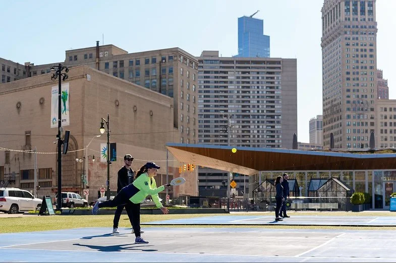 People playing pickleball on an outdoor court in an urban area with tall buildings and cityscape in the background.