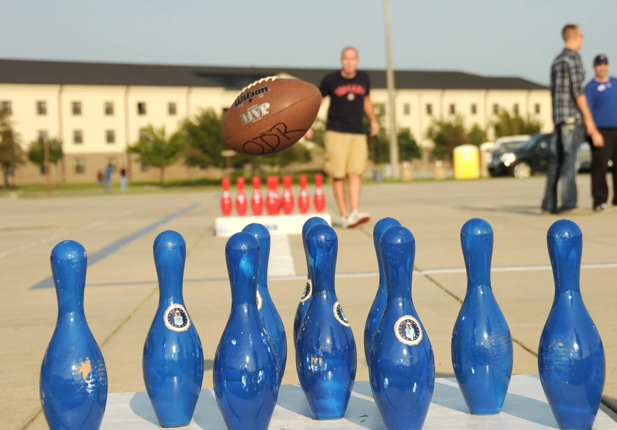 Blue bowling pins set up on a sidewalk with a brown football in mid-air heading towards them, with people and a building in the background.