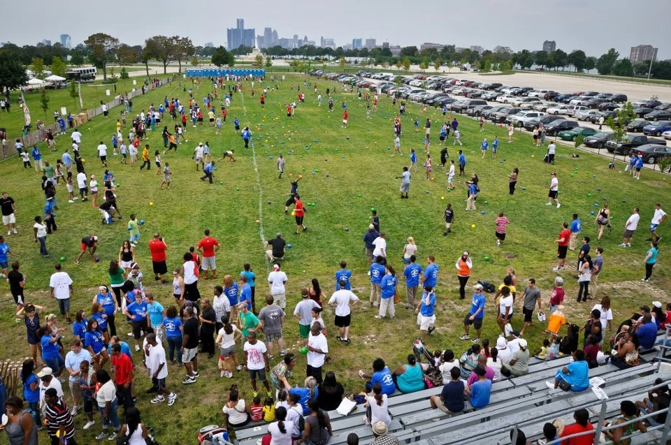 Crowd of people participating in an outdoor event on a grassy field, with some in blue shirts and others playing games with yellow and green balls, overlooking a parking lot and city skyline.