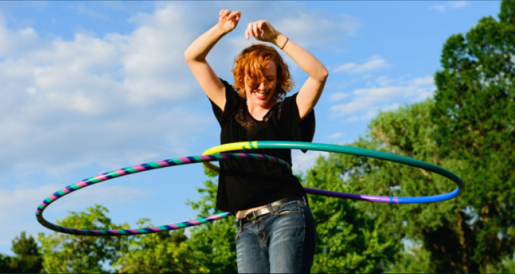 A young woman with curly red hair hooping outdoors against a blue sky with clouds and green trees.