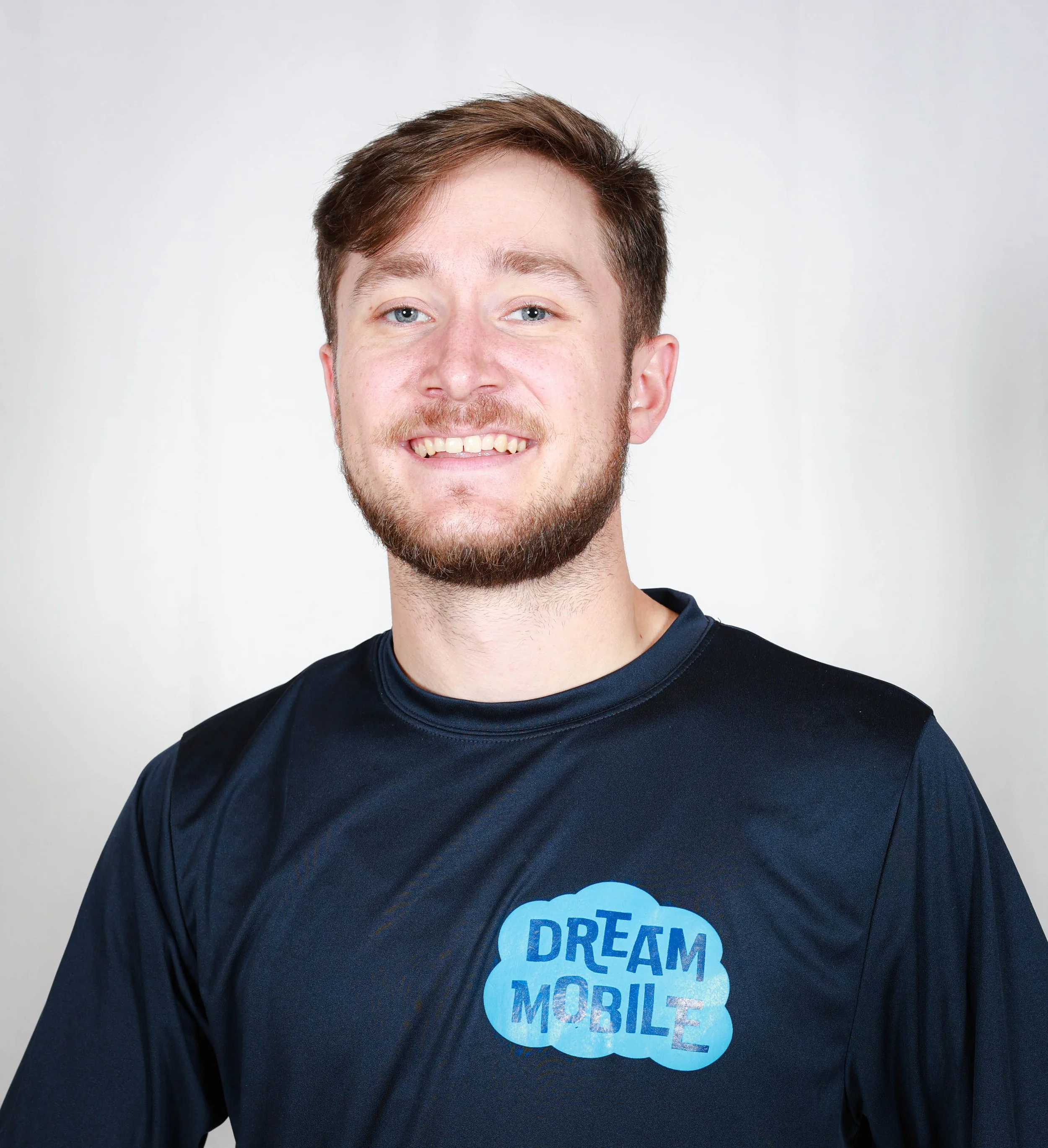 A young man with light skin, brown hair, and a beard smiling, wearing a black t-shirt with a blue cloud logo that says 'DREAM MOBILE'.