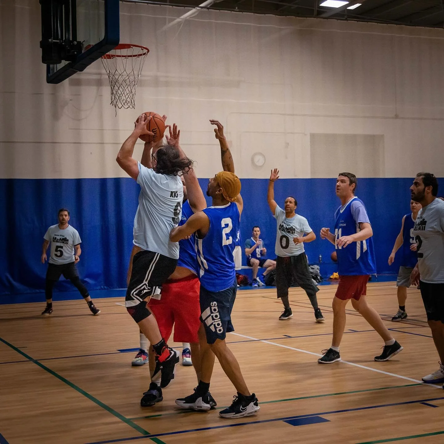 People playing basketball indoors, one player jumping to shoot or block the ball near the hoop, with others defending and watching, inside a basketball court.