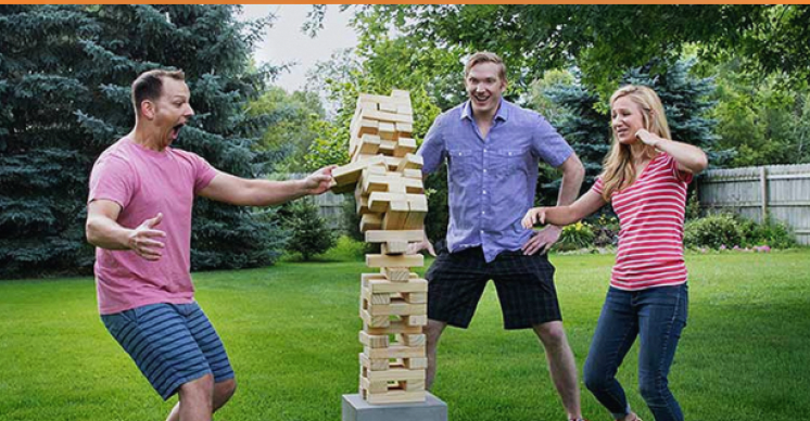 Three people playing giant Jenga outside in a backyard with green grass, trees, and a wooden fence, enjoying a fun game.