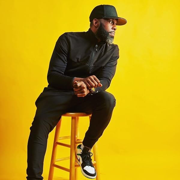 A man with a beard, wearing a black jacket, black sweatpants, black cap, and black and white sneakers, sitting on a yellow stool against a yellow background.