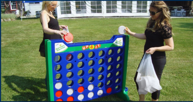 Two women playing giant outdoor Connect Four game on grass, with white building and trees in background.