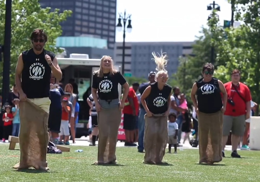 People participating in a sack race at an outdoor event in a park with a crowd watching.