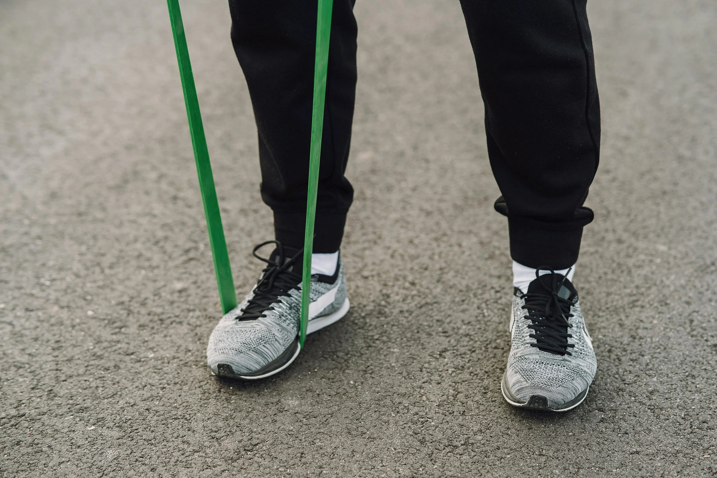 Person wearing athletic shoes and black pants, standing on a paved surface, holding green resistance bands.