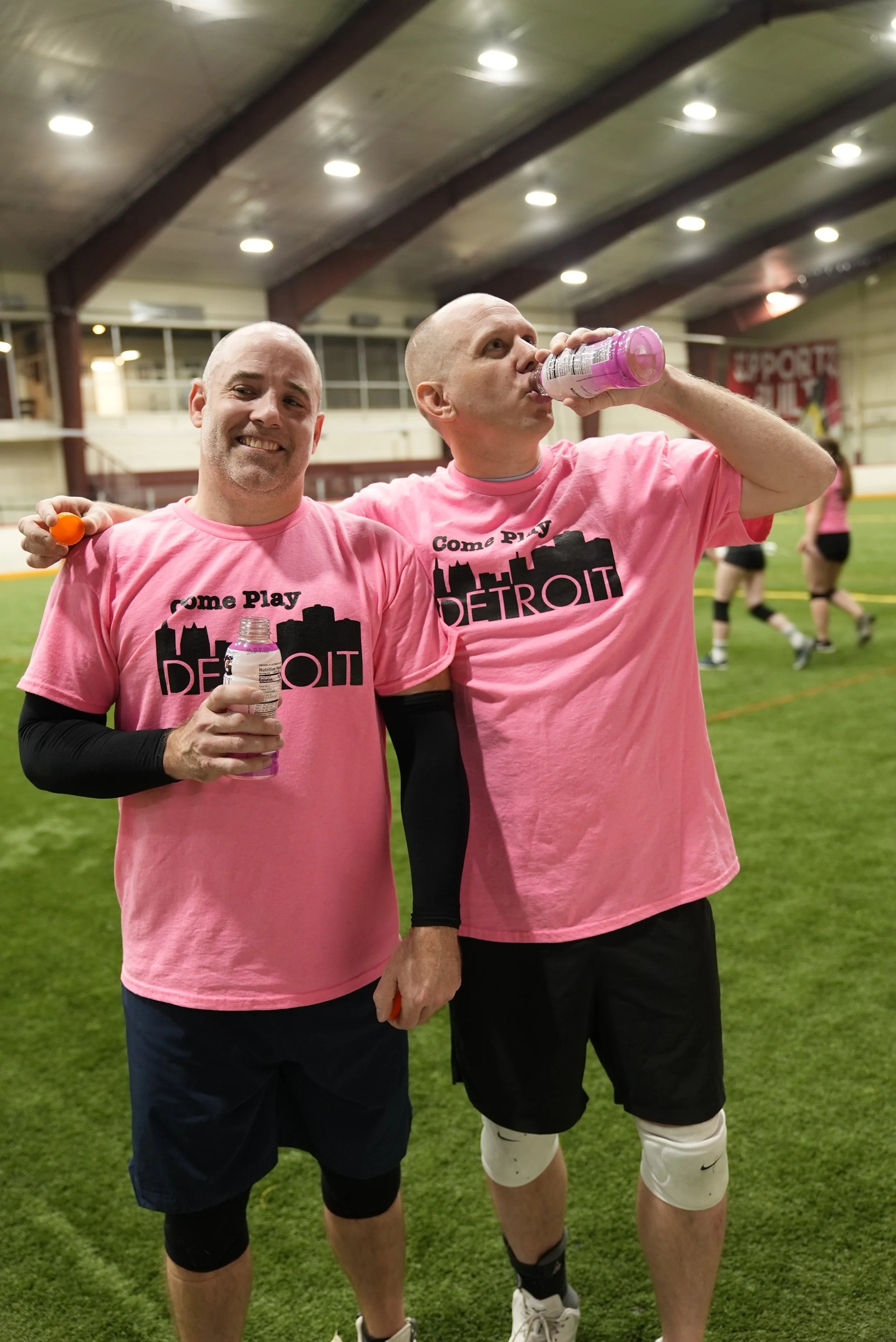 Two men in pink T-shirts with 'Come Play Detroit' logo standing on an indoor sports field, one smiling and the other drinking water, with other players in the background.