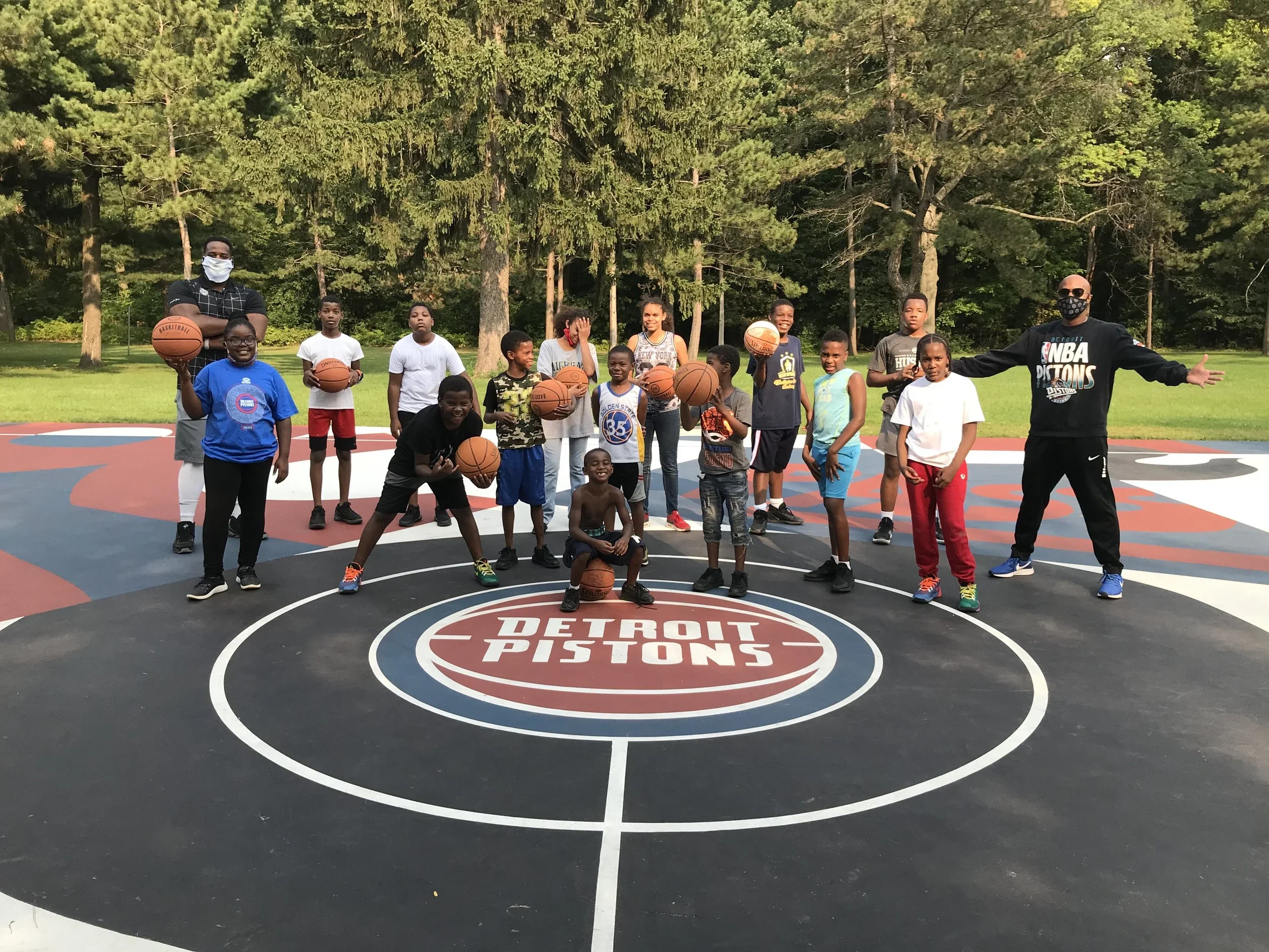 A group of children and two adults on a Detroit Pistons themed basketball court outdoors, some holding basketballs, among trees and greenery.