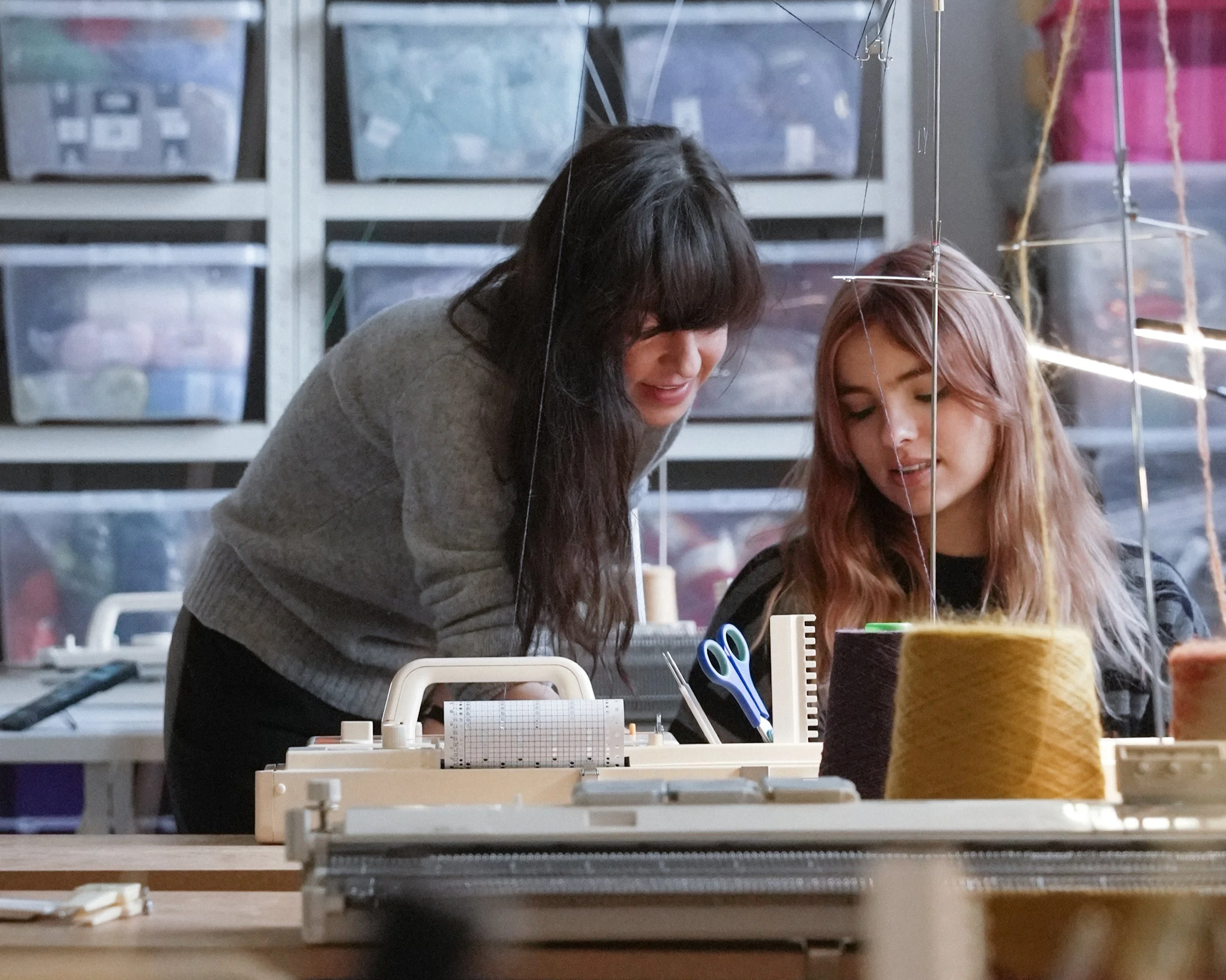 Two women working with yarn and knitting on a vintage knitting machine in a textile studio with shelves filled with clear containers full of colourful yarn in the background.