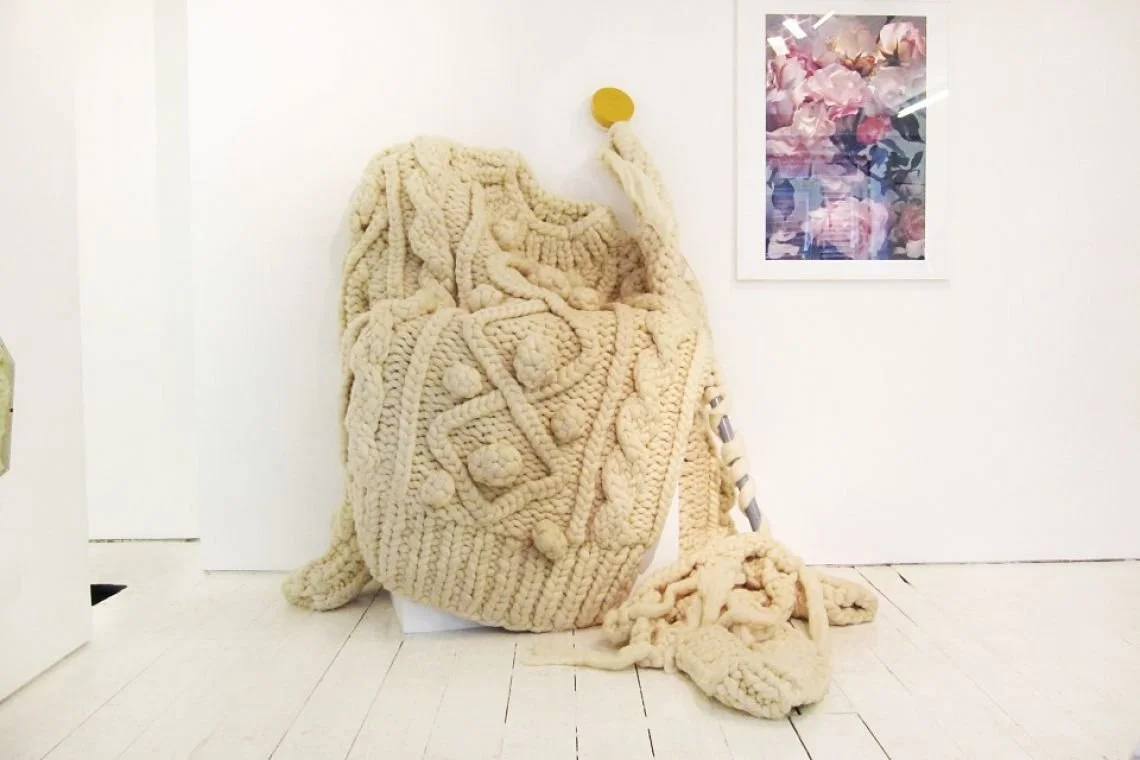 Large knitted art piece resembling a cable knit sweater with textured patterns, standing upright against a white wall, with a colourful abstract painting to the right.