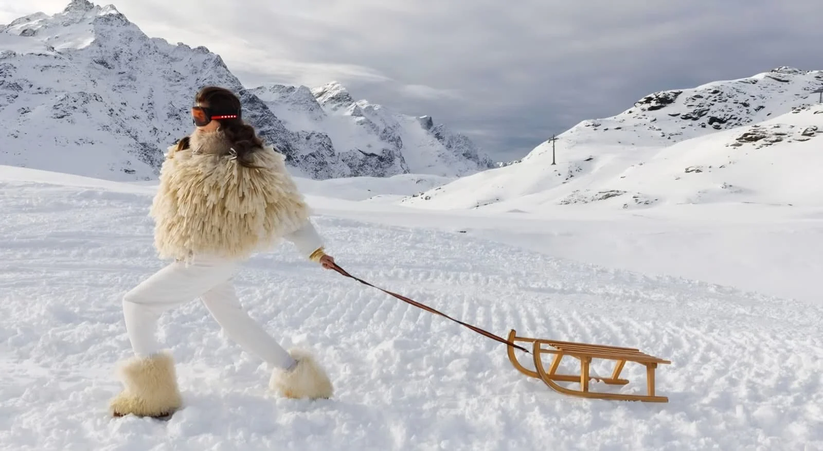 Person dressed in high-fashion winter clothing and furry boots, pulling a wooden sledge in a snowy mountain landscape.