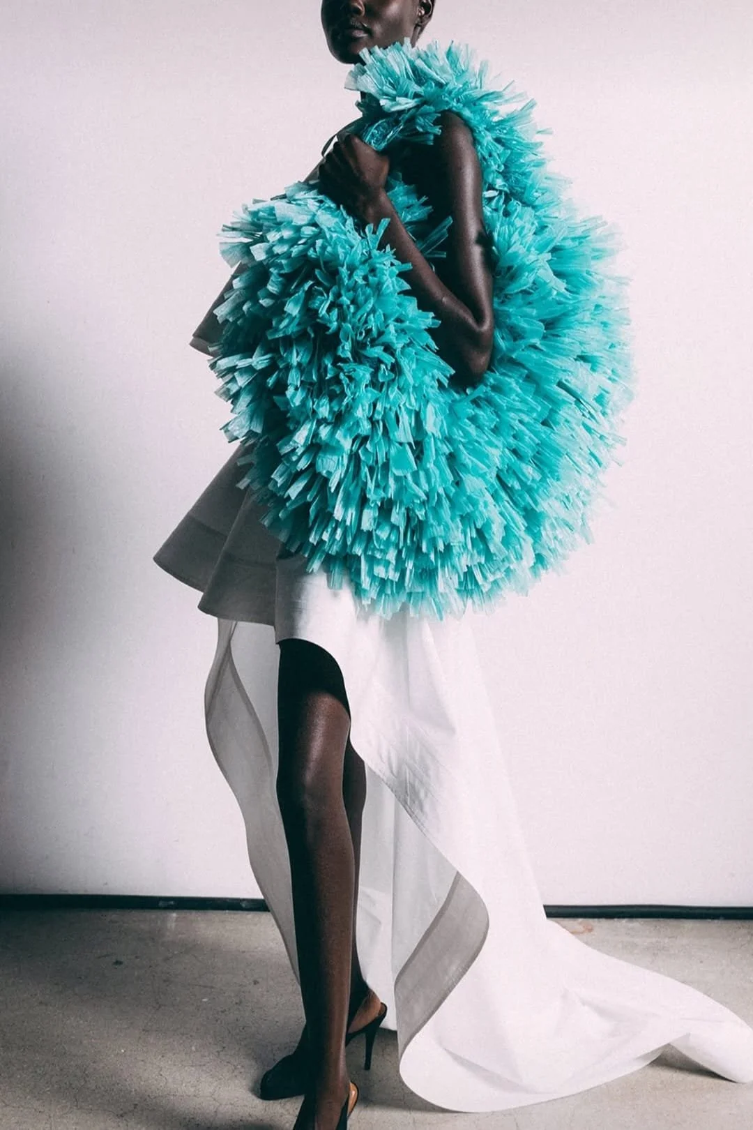 A black model wearing a large, fluffy blue handbag made of layered, crinkled fabric, standing against a plain white wall, wearing a thin white dress.