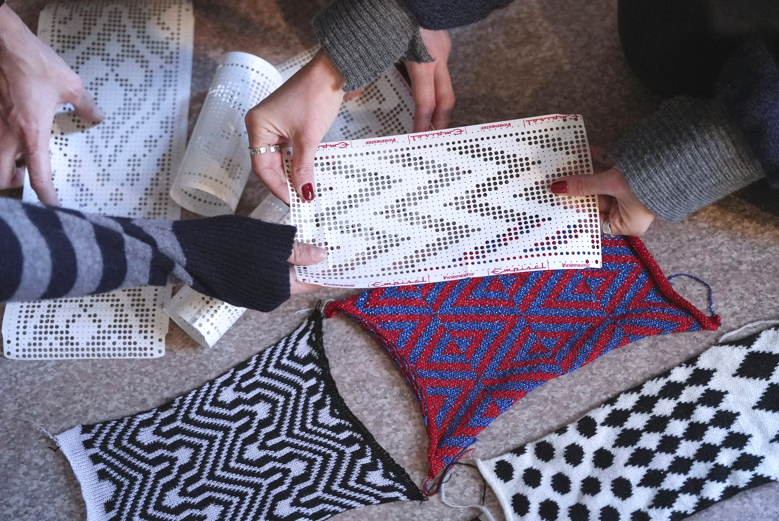 People are working on a project using punch cards with various geometric patterns. The individuals' hands are visible, some holding the cards and others working with textiles. There are colourful, patterned pieces of fabric or yarn nearby.