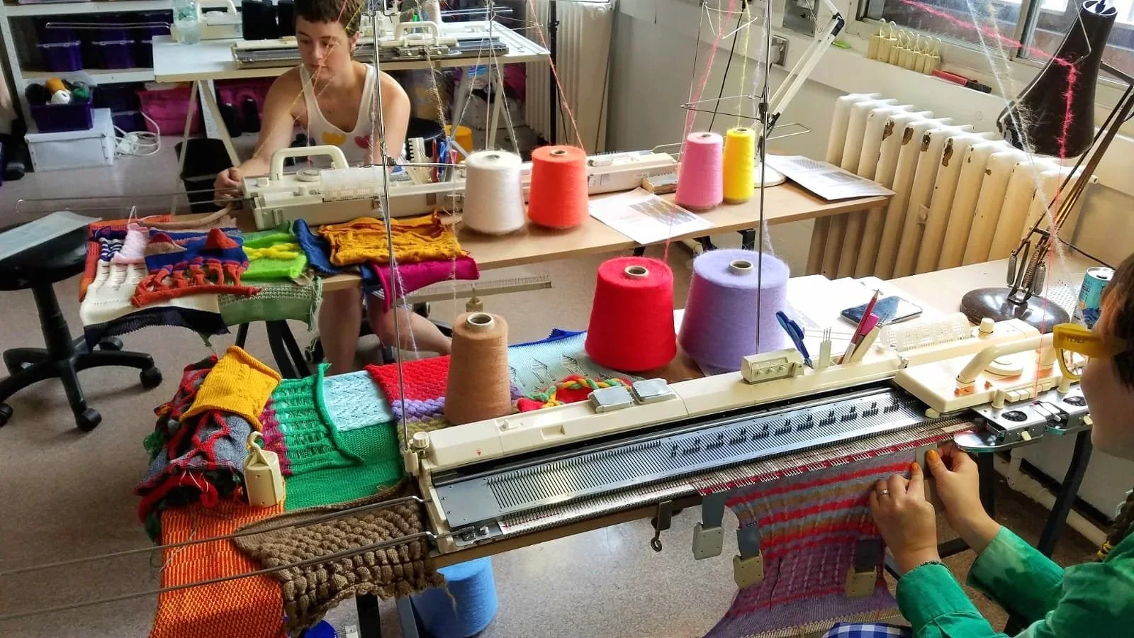 People are working on knitting machines in a bright indoor space. Spools of colourful yarn and knitted items are on the tables.