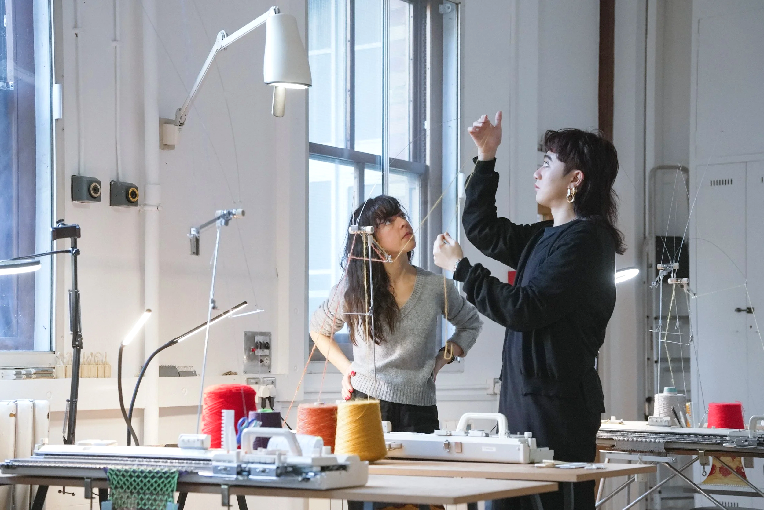 Two women working on a textile project in a well-lit workshop with large windows, vintage knitting machines, and colourful spools of thread.